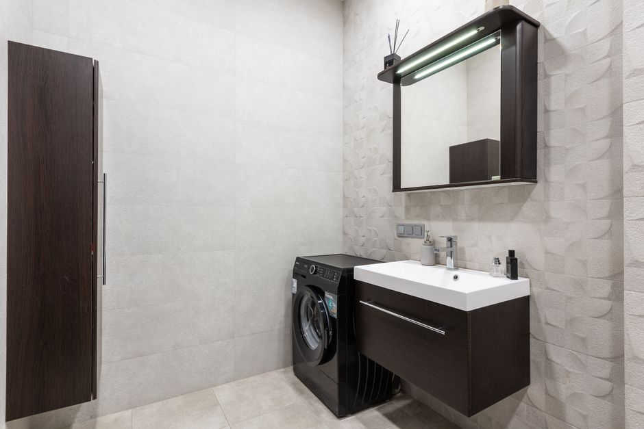 Modern black washing machine placed at wall near sink with dispenser and mirror in stylish light bathroom with brown cupboard